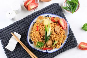 Mie goreng or Stir Fried Egg Noodles is Traditional Indonesian Food with Chicken, Egg, Tomato, Onion, Vegetable and Sweet Soy Sauce in White Plate Served on Wooden White Table. Top View.