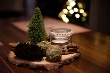 Stroik świąteczny z choineczką, świeczką, cynamonem i szyszkami na stoliku kawowym z mieniącą się choinką w tle.