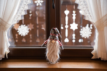 Świątecznie przystrojone okno, krasnal z długą białą brodą na parapecie. Do szyb przyklejone białe śnieżynki.