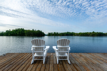 White Adirondack chair on a wooden dock facing the blue water of a lake in Ontario Canada. Cottages...