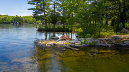 Brown Adirondack chairs on a rock formation facing a lake in Muskoka, Ontario Canada. Across the...