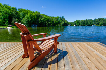 A brown Adirondack chair on a wooden pier facing a lake in Muskoka, Ontario Canada. Across the calm...