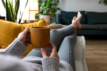 POV of young woman relaxing at home with cup of coffee lying on couch.