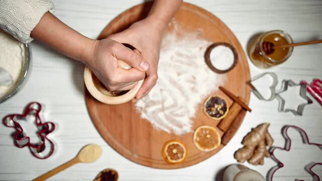 Top view. Female hands grinding spices and ingredients in marble mortar. On the kitchen surface, there is wooden board sprinkled with flour, dried orange slices, molds for cutting gingerbread cookies