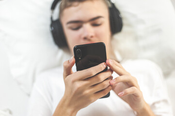 Teenager lies in bed with headphones and watches videos or listens to music on a smartphone Digital addiction Online communication and entertainment. Focus on the hand with the phone Selective focus.