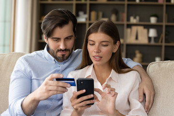 Focused millennial couple of consumers using banking app on mobile phone, credit card for online payment, transaction, transfer, money order, shopping on internet, paying bills, entering data