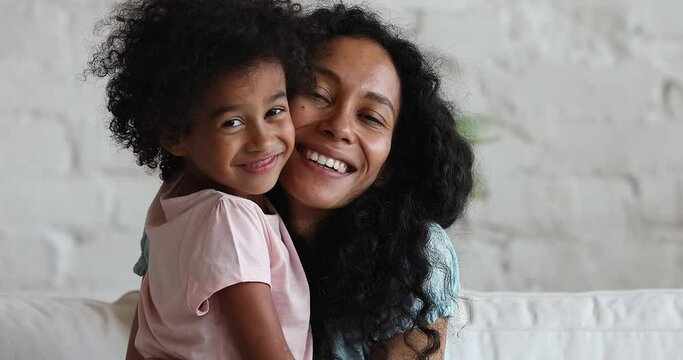 Close up portrait African mother her cute little 5s daughter. Woman and kid sit on couch smile look at camera cuddling enjoy sweet moment together feeling unconditional love. Happy Mothers Day concept