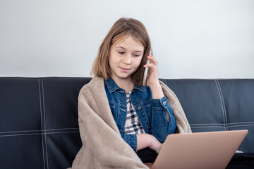 A teenage girl is sitting on the couch at a laptop and talking on the phone.