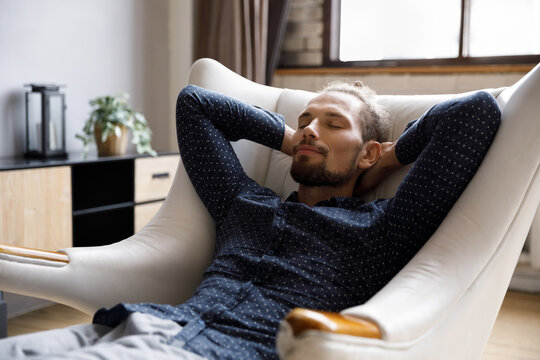Calm happy millennial guy relaxing in pale comfortable armchair at home, resting with sleepy face, closed eyes, breathing fresh air deeply for stress reduce, calmness, serenity, meditation