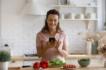 Happy smiling girl preparing salad from fresh vegetables, cooking in home kitchen, reading recipe on food blog, chatting online, texting message, making call on mobile phone at table