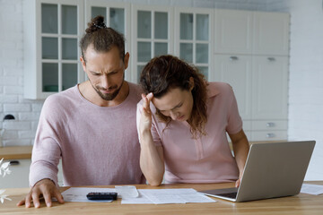 Frustrated concerned millennial married couple calculating overspend budget, doing paperwork job at...
