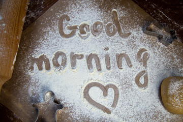 On the baking paper on the flour is written Good morning and heart, against the background of dough, rolling pins and cookie cutters. High quality photo