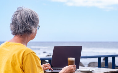 Back view of mature woman sitting at coffee table drinking special coffee using laptop computer. Horizon over water