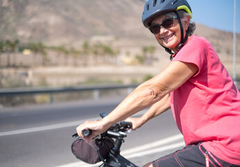 Fototapeta na wymiar Portrait of happy mature woman, wearing protective helmet, on her bike in the sunny road. Healthy and active retirement concept