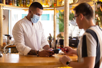Young Latin American waiter with face mask charging from the bar to a customer by contactless payment.