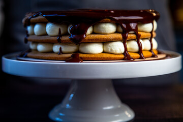 A cake with brown honey cakes, white cream and poured on top with dark chocolate, which flows down the sides, stands on a white stand on a dark background. High quality photo