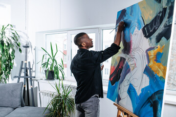 Immersed black man painting on a big canvas inside of his apartment