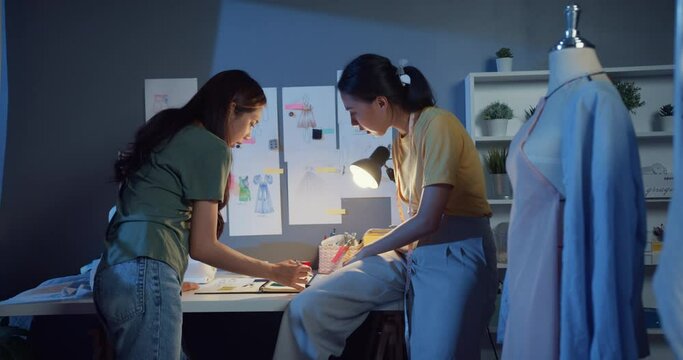Two professional Asia cheerful lady fashion designer teamwork with casual overtime work discuss meeting on table share idea create dress collection at night. Lady tailor partner start up business.