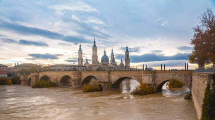 Fototapeta premium Stone bridge next to the Basilica De Nuestra Señora del Pilar on the Ebro river in the city of Zaragoza at sunset, Aragon. Spain