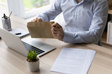 Close up cropped view businessman sit at workplace desk takes letter or bill from envelope, sorting...