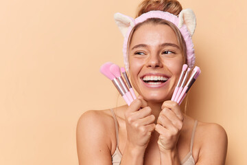 Happy sincere European woman with healthy clean skin holds makeup brushes near face smiles broadly undergoes beauty treatments poses bare shoulders against brown background applies cosmetics