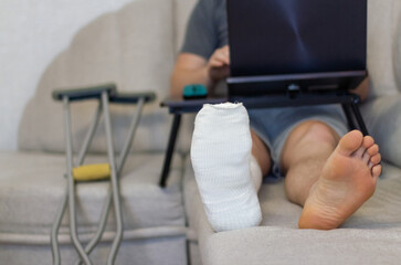 Man is lying on sofa and works at the computer with bandage on leg.