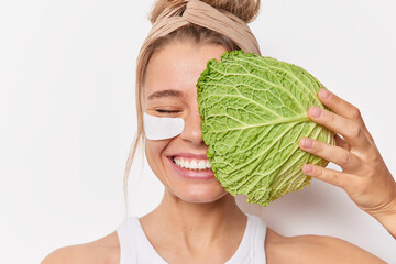 Happy sincere young woman takes care of skin covers face with cabbage leaf applies patches under...