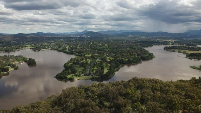 Aerial drone shot lifting high above the tree tops over a beautiful landscape with a lake, small city, forests and mountains. In the background there is a storm with rain falling. Filmed in Canberra. 