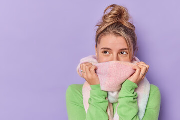 Thoughtful female model feels cold mouth with scarf tries to warm looks away poses against purple background blank copy space for your promotional content. People and winter clothes concept.