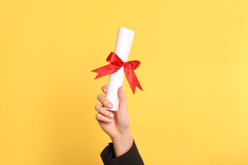 Student holding rolled diploma with red ribbon on yellow background, closeup