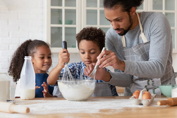 Loving African dad teach kids to cook in kitchen, hold beaters mixing ingredients in bowl preparing...