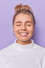 Vertical shot of pleased attractive woman smiles toothily keeps eyes closed imagines something wears white jumper isolated over purple background. People happy emotions and feelings concept.