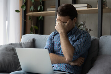 Man sit on sofa with laptop cover face with hand got bad news feels disappointed, looks distressed with awful e-mail, having problems, miss appointment, confused by mistake or error, troubles concept