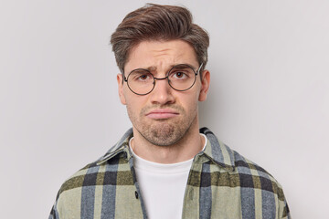 Obraz premium Portrait of sad unhappy man frowns face upset with bad news he received wears round spectacles and casual checkered shirt isolated over white background feels reluctant. Negative emotions concept