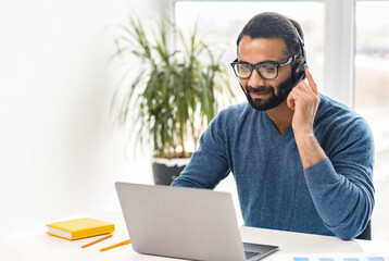 Smiling Indian office worker in glasses and headset looking at laptop screen and talking online, positive ethnic man holding video call, working in customer service department, explaining something