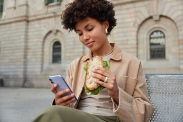 Pleased beautiful curly haired woman eats sandwich and uses mobile phone for surfing internet or...