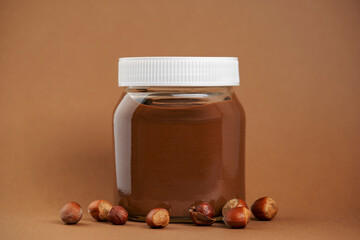 Chocolate spread or nougat cream with hazelnuts in glass jar on brown background. 