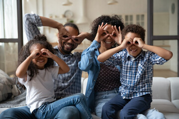 Happy African family with little kids make funny faces showing binoculars, circles shape with fingers smile look at camera, enjoy funny games sit on sofa at home. Vision check up, eyewear shop concept
