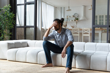 Obraz premium Young African guy sit on sofa at home touch head looks desperate, suffers from headache, goes through divorce feel mental pain, depressed due to hard life situation, break up, personal trouble concept