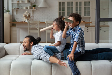 African dad lying on couch spread arms like plane wings look at distance, joyful kids sit on daddy...