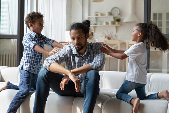 Hyperactive African little children want to play with tired annoyed father sit on sofa feels exhausted to spend playtime with kids. Upbringing difficulties, need rest, divorced helpless parent concept