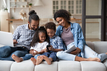 African couple with little children sit on sofa with diverse devices, hold tablet, laptop, smartphone. Mobile application usage, bad habit, free time use modern tech, gen addicted with gadgets concept