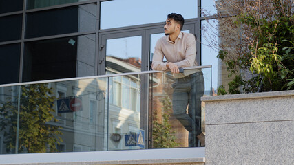 Confident young hispanic business man standing at railing modern skyscraper office building looking...