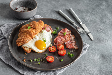Croissant sandwich with fried egg and bacon. Gray grunge background. Delicious appetizing breakfast. Side view, selective focus.