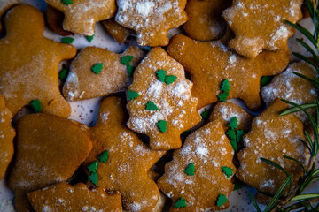 Cookies are on a plate of different shapes, in the foreground there is a cookie in the form of a Christmas tree, decorated with powdered sugar like snow and small green Christmas trees against the