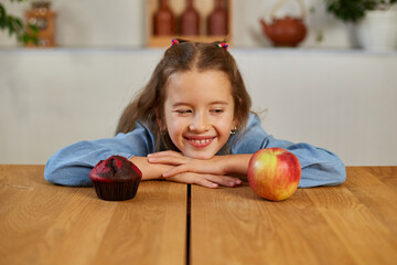Fototapeta premium Little girl looking on sweet cake and fresh red apple and choosing in kitchen at home