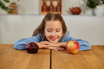 Fototapeta premium Little girl looking on sweet cake and fresh red apple and choosing in kitchen at home