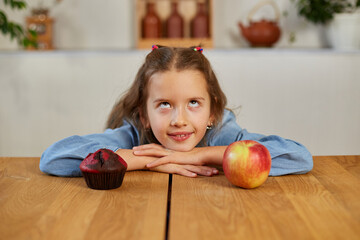 Fototapeta premium Little girl looking on sweet cake and fresh red apple and choosing in kitchen at home