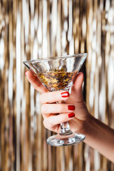 Woman hand is holding martini glass with confetti in the middle of Golden foil tinsel strips. Festive background for christmas, new year, holidays or birthday celebration card.