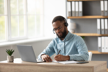 Busy serious millennial african american bearded guy in glasses and headset working on laptop in...